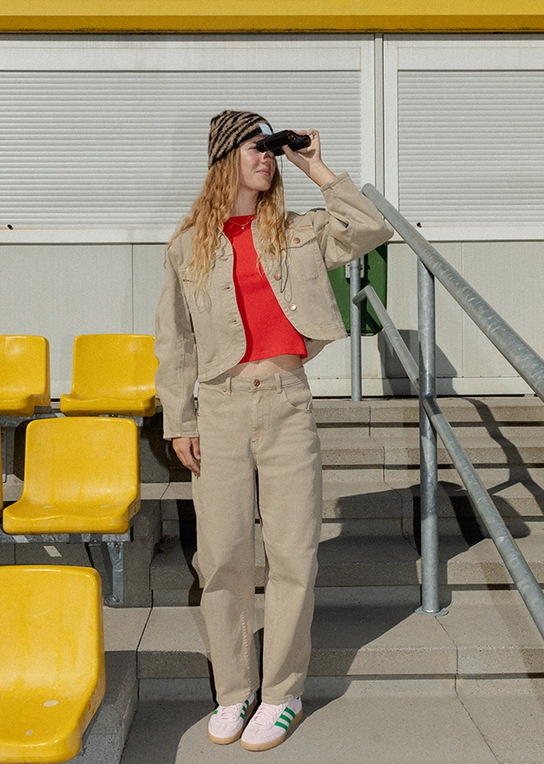 Nika steht auf einer Stadiontribüne und schaut durch ein Fernglas. Sie trägt ein rotfarbenes T-Shirt, eine beigefarbene Jacke mit passender Hose und eine Mütze mit braun-schwarzem Tigermuster; Zur abgebildeten Hose gehen