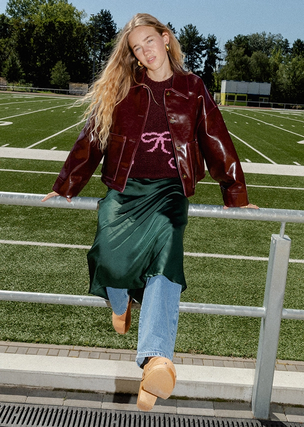 Nika steht in einem Footballstadion an ein Geländer gelehnt. Sie trägt einen langen dunkelgrünen Satinrock über einer blauen Jeans und dazu eine bordeauxrote Lederjacke mit einem dunklen Strickpullover darunter; Zum abgebildeten Rock gehen
