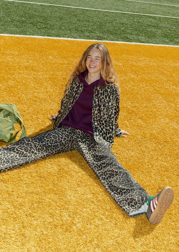 Nika sitzt auf einem gelben Sitz auf einer Stadiontribüne. Sie trägt eine cremeweiße Bluse mit einer gestreiften Strickweste in Orange und Lila darüber. Dazu trägt sie eine blaue Jeans und goldenen Schmuck; Zur abgebildeten Weste gehen