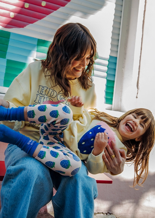 Julia sitzt lachend auf einer roten Bank und hat ein lachendes Mädchen auf dem Schoß. Julia trägt einen hellen gelben Sweater mit ?HDGDL?-Schriftzug und eine blaue Jeans. Das kleine Mädchen trägt einen hellgelben Sweater mit einer großen, blauen Erdbeere. Dazu trägt sie eine gerippte Leggins mit blauem Erdbeermuster und blauen Socken; Zur abgebildeten Kids-Leggins gehen