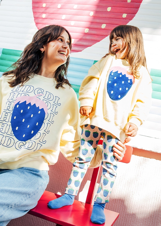 Julia sitzt auf einer roten Bank und neben ihr steht ein kleines Mädchen auf der Bank. Beide sehen sich lachend in die Augen. Julia trägt einen hellgelben Sweater mit großer blauer Erdbeere und ?HDGDL?-Schriftzug vorne drauf. Das Mädchen trägt einen hellgelben Sweater mit großem, blauem Erdbeermotiv bestickt. Dazu trägt sie eine helle Leggins, die mit vielen kleinen Erdbeeren bedruckt ist und passend blaue Socken; Zum abgebildeten Damen-Sweater gehen