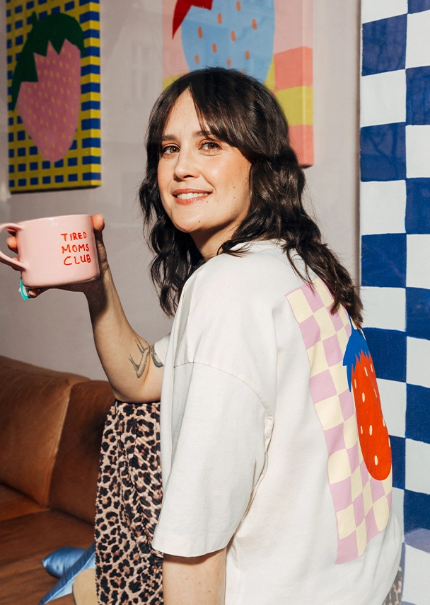 Julia sitzt entspannt auf einem Sofa und hält lächelnd eine rosafarbene Tasse mit rotem ?Tired Moms Club?-Schriftzug und breitem Henkel in der Hand. Sie trägt ein cremefarbenes T-Shirt mit großem Backprint einer rot-blauen Erdbeere auf einem rosa-gelben Schachbrettmusterhintergrund und eine Leopardenprinthose; Zur abgebildeten Tasse gehen