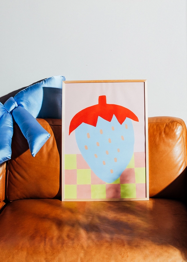 Ein eingerahmtes Poster steht auf einem braunen Ledersofa. Auf dem Poster ist eine große hellblau-rote Erdbeere auf einem rosa-gelb gemusterten Hintergrund abgebildet; Zum abgebildeten Poster gehen