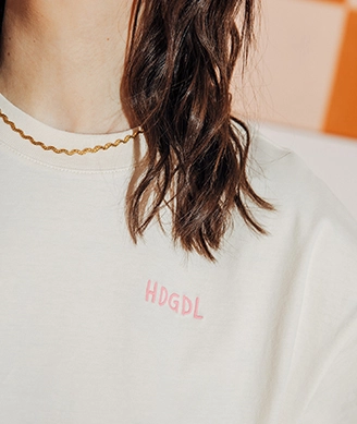 Beim Bild liegt der Fokus auf Julias Oberkörper. Sie trägt ein cremefarbenes T-Shirt mit rosafarbenem ?HDGDL?-Schriftzug auf der linken Brust. Dazu trägt sie eine goldene Halskette. Auf dem zweiten Bild sitzt Ani mit angewinkelten Beinen auf dem Boden und lächelt. Sie trägt ein hellgelbes T-shirt mit großem Backprint einer rot-blauen Erdbeere auf einem rosa-gelben Schachbrettmusterhintergrund und dazu eine Leopardenprinthose mit silbernen Sneakern; Zum abgebildeten T-Shirt gehen