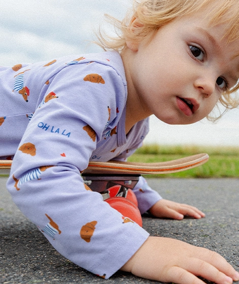Ein kleines Kind liegt mit dem Bau auf einem Skateboard und trägt einen lilafarbenen Body mit kleinen, bunten Croissants und Dackeln bedruckt; Zum abgebildeten Body gehen