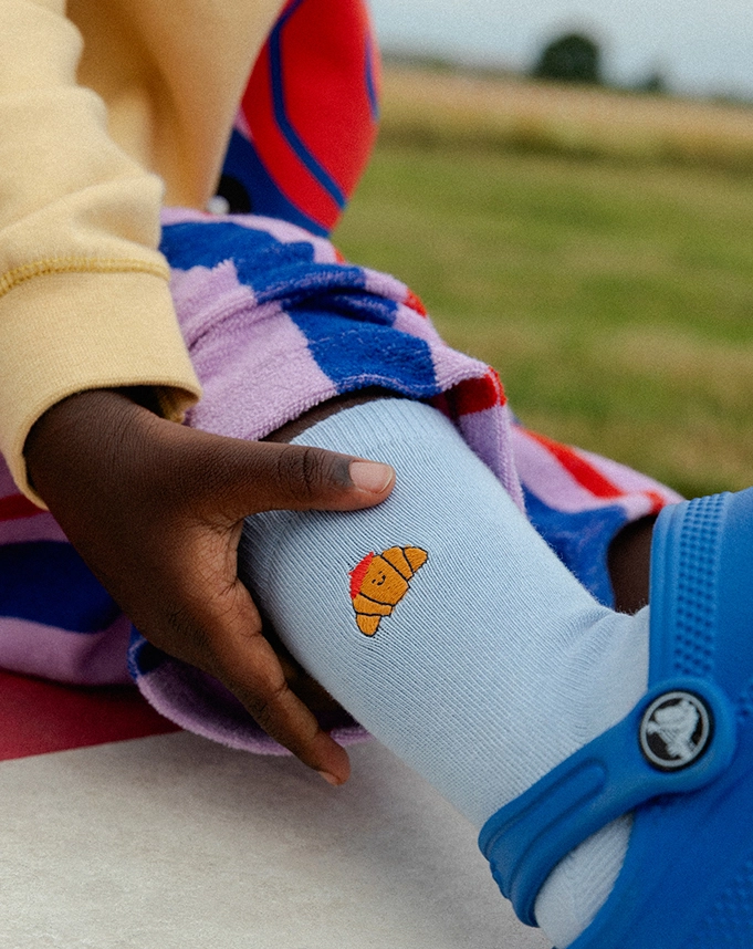 Ein Kind trägt eine lilafarbene Frotteehose mit blauen und roten Streifen und eine Hellblaue Socke mit niedlicher Croissant-Stickerei am Bund; Zur abgebildeten Socke gehen