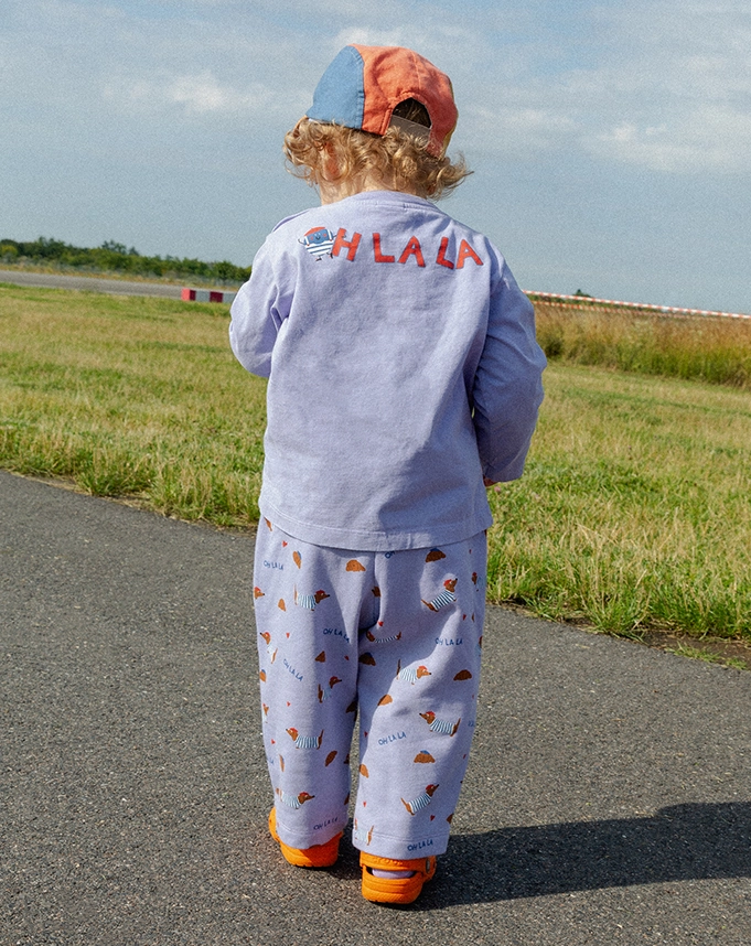 Ein Kind steht auf einem Weg vor einer Rasenfläche und trägt ein lilafarbenes Longsleeve mit rotem ?OH LA LA?-Backprint und eine farblich dazu passende, mit kleinen Dackeln und Croissants bedruckte Sweathose. Auf dem Kopf trägt es eine bunte Cap; Zum abgebildeten Longsleeve gehen