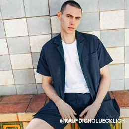 Auf dem Bild sieht man einen sitzenden Mann vor einer Fliesenwand. Er trägt ein offenes kurzärmliges Hemd in blau und eine passende Shorts. Dazu kombiniert er ein weißes T-Shirt. Das Bild ist vom Instagram Account von @KAUFDICHGLUECKLICH; Zum abgebildeten Hemd gehen.
