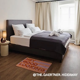 Ein großes Bett steht in einem Raum vor einem Fenster und auf dem Nachttisch leuchtet eine Lampe. Vor dem Bett liegt ein kleiner Teppich mit lila- und orangefarbenen Streifen und dem Schriftzug 