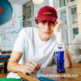 Kim sitzt an einem Holztisch und hat den Kopf auf die Hand gestützt. Er trägt ein weißes T-Shirt und eine dunkelrote Cap mit weißem ?TUTTO BENE?-Schriftzug darauf; Zur abgebildeten Cap gehen