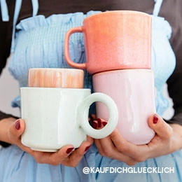 Eine Frau trägt ein hellblaues, gerafftes Top, welches sie über einem schwarzen Longsleeve kombiniert hat. Vor ihrem Oberkörper hält sie gestapelte und verschiedenfarbige Tassen in unterschiedlichen Formen in den Händen; Zur abgebildeten Tasse gehen