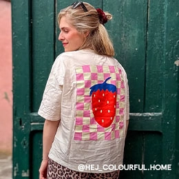 Katha steht vor einer grünen Tür und schaut halb über ihre Schulter. Sie trägt ein Cremefarbenes T-Shirt mit großem, buntem Erdbeer-Backprint und dazu eine Hose mit Leopardenmuster; Zum abgebildeten T-Shirt gehen