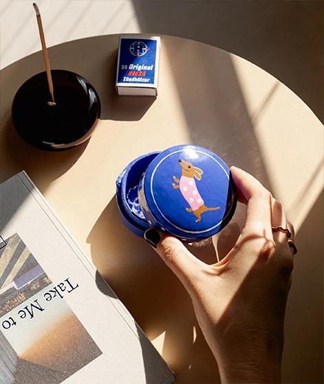 Auf einem beigen Beistelltisch steht ein dunkelroter Räucherhalter aus Glas mit Räucherstäbchen. Daneben liegen Streichhölzer, ein Buch und eine Schmuckdose. Zu unserer Home-Kollektion.