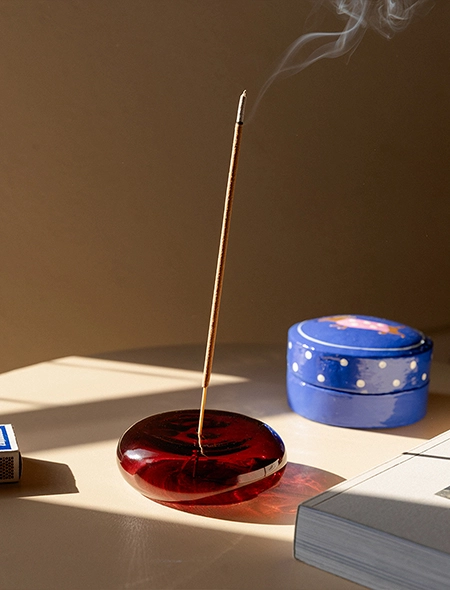 Auf einem beigen Beistelltisch steht ein dunkelroter Räucherhalter aus Glas mit Räucherstäbchen im Vordergrund. Daneben liegen Streichhölzer, ein Buch und eine Schmuckdose. Zu unseren Neuheiten.
